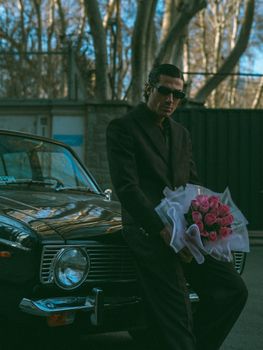 A stylish man in sunglasses leans on a vintage car holding pink roses, exuding a mysterious charm.