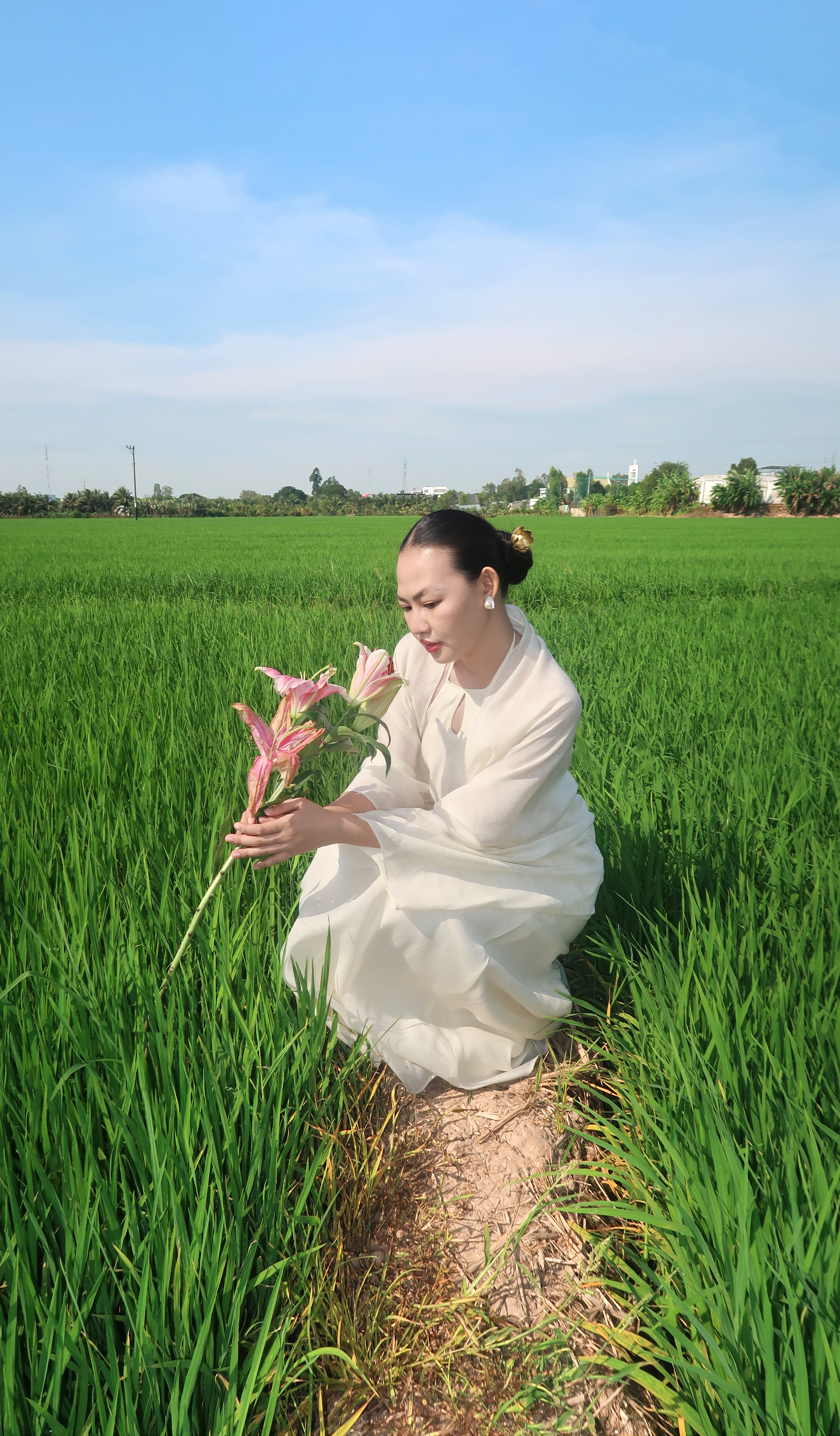 A serene woman kneels in a green field, holding pink flowers under a clear blue sky.
