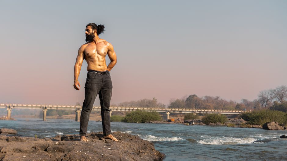 A shirtless muscular man standing confidently on rocks beside a serene river with a bridge in the background.