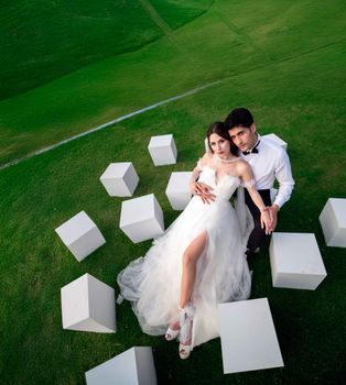 Bride and groom in a stylish pose outdoors surrounded by white cubes on green grass.