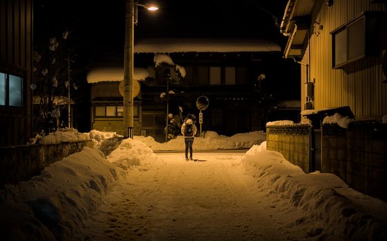 A lone figure walks a snow-covered street under a warm streetlight in Eiheiji, Japan.