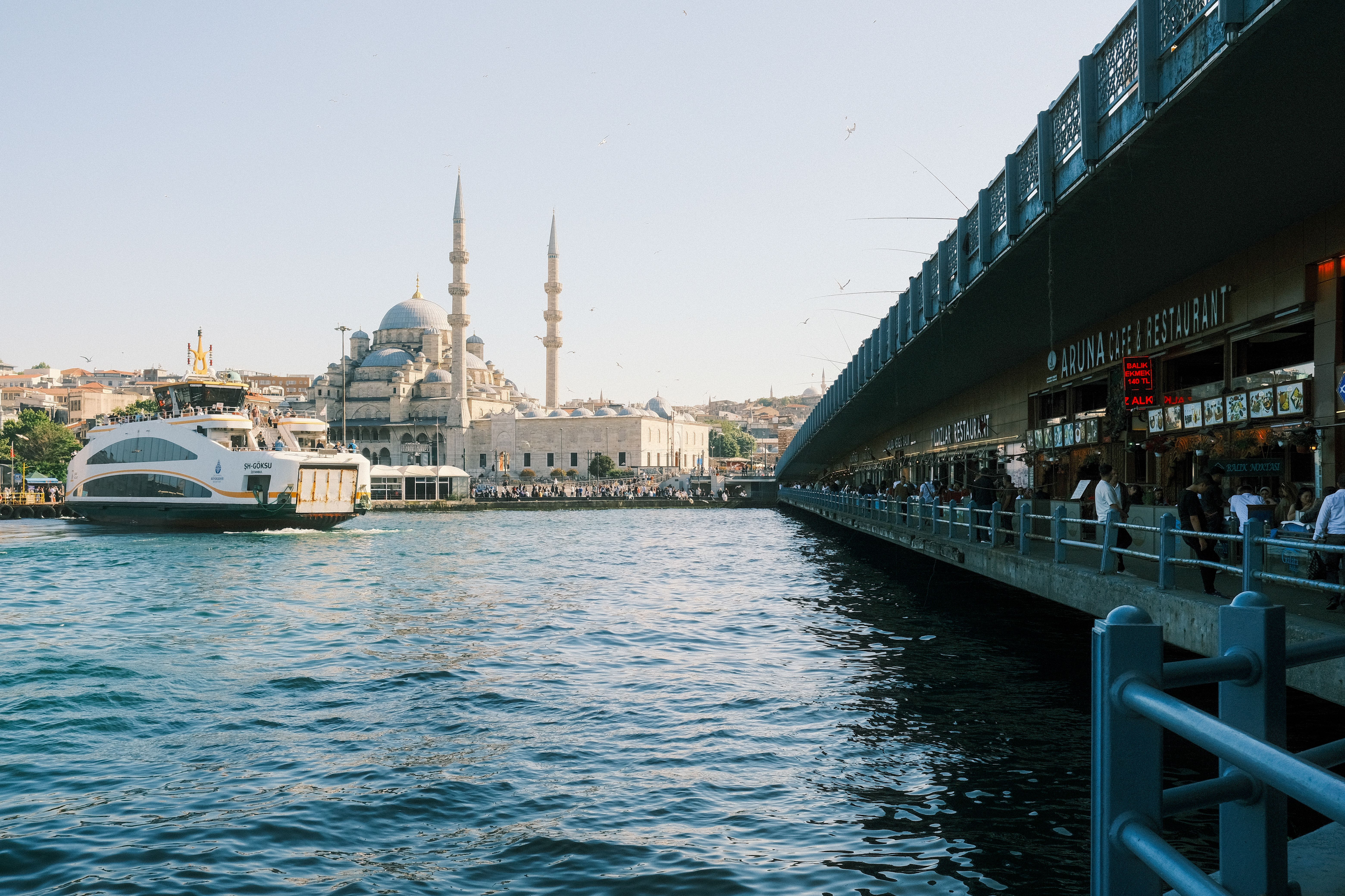 A white passenger ferry on blue water beside a long bridge with restaurants along its lower deck, and a mosque with two minarets on the far shore