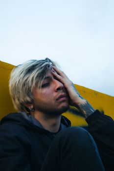 A young adult with contemplative expression sitting against a vibrant yellow wall outdoors.