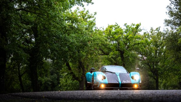 Sleek sports car with blue accents drives through a green forest road, headlights on, during the daytime.