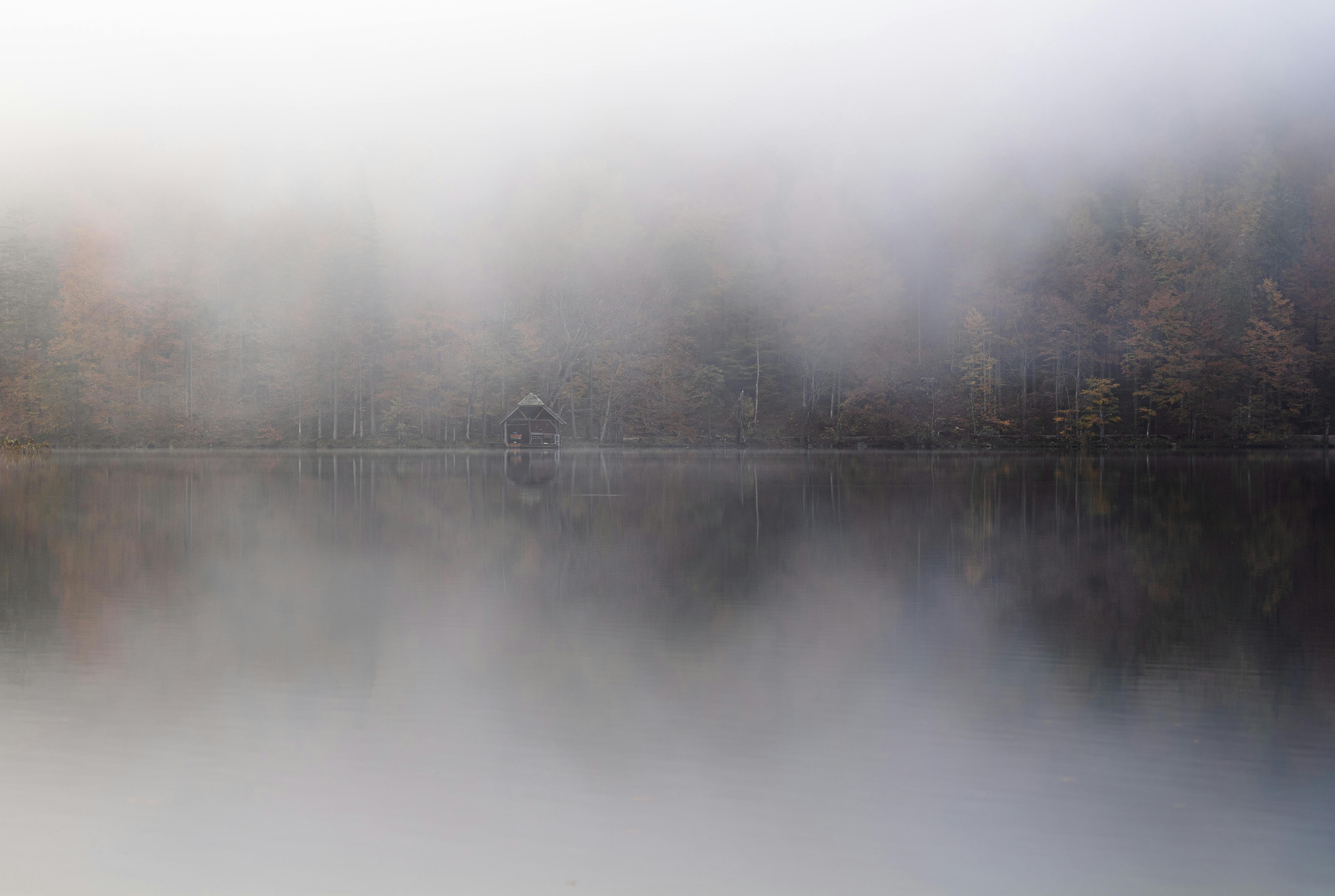 A tranquil lake with a cabin partially obscured by fog and surrounded by autumn foliage.