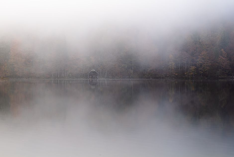 A tranquil lake with a cabin partially obscured by fog and surrounded by autumn foliage.