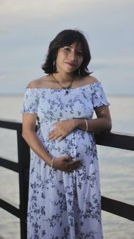 A pregnant woman in a floral dress standing by the seaside at sunset, expressing serenity.