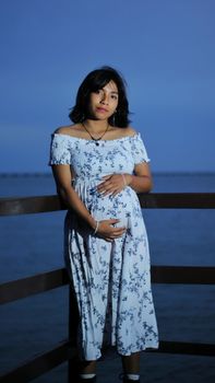 A serene portrait of a pregnant woman in a floral dress by the ocean at twilight.