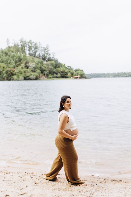 Free Expectant mother in casual attire walking by a serene beach with lush greenery. Stock Photo