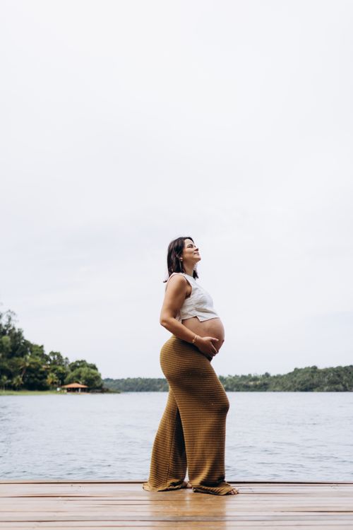 Free Side view of a pregnant woman wearing a white top and brown pants enjoying a peaceful lakeside setting. Stock Photo