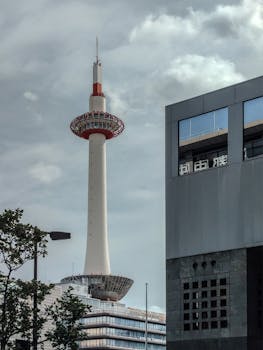 View of Kyoto Tower with modern architecture in the foreground under a cloudy sky.