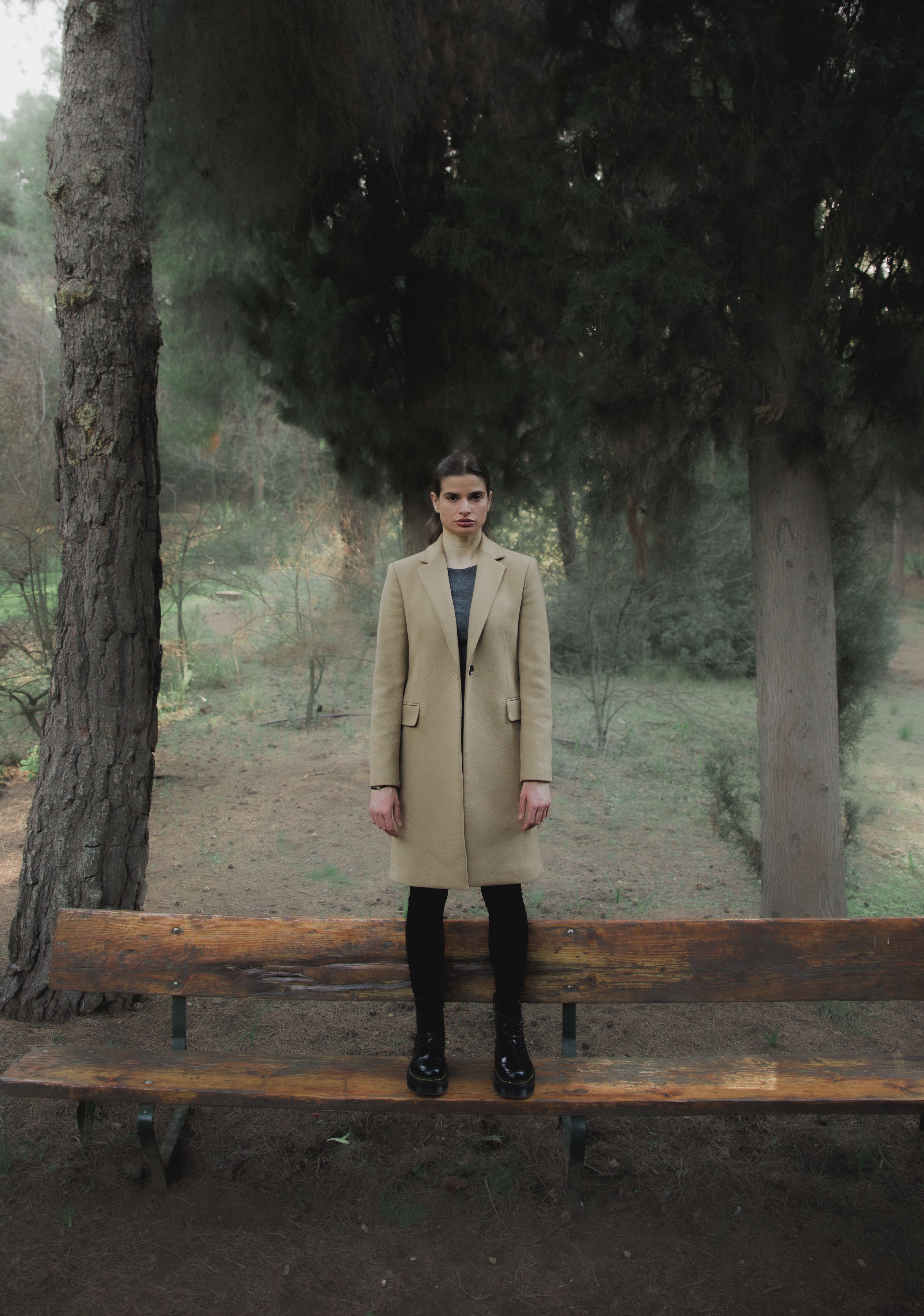 A young woman stands on a bench in a serene, wooded park wearing a tan coat.