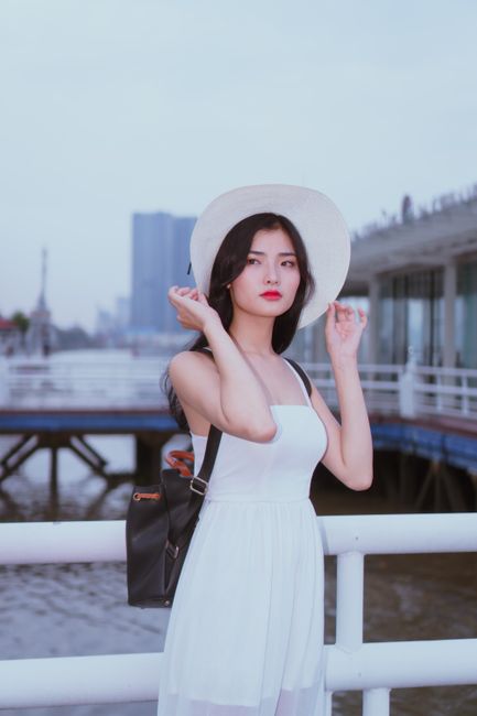 Elegant woman in white summer dress and hat posing by the waterfront, exuding fashion and style.