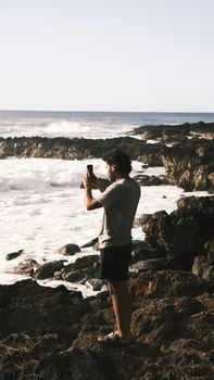 A man takes a photo of ocean waves on a rocky coastline, capturing nature's power.