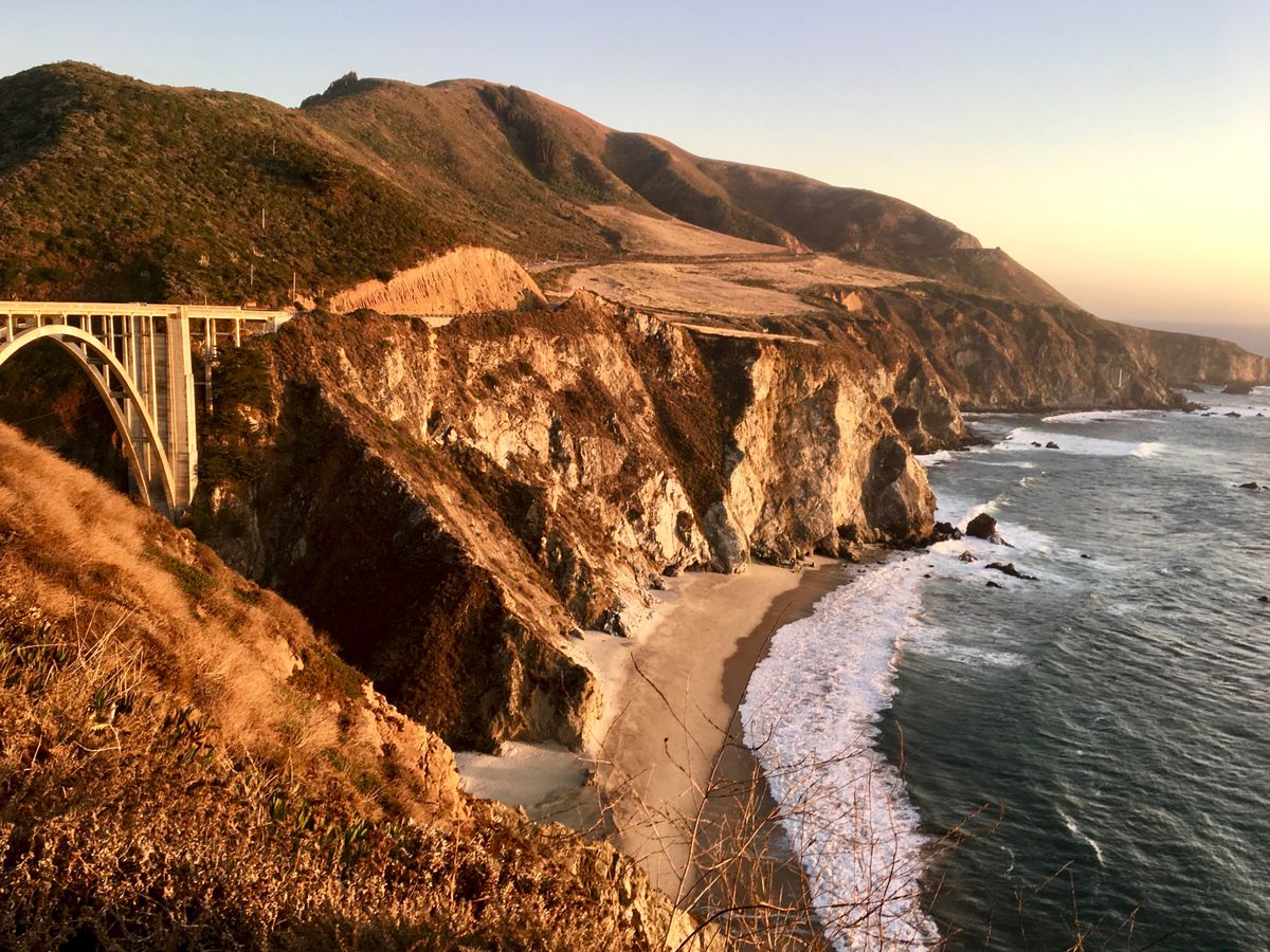 Bixby Creek Bridge at golden hour from the north turnout, with cliffs dropping to a sandy cove and Highway 1 curving along the hills behind it.