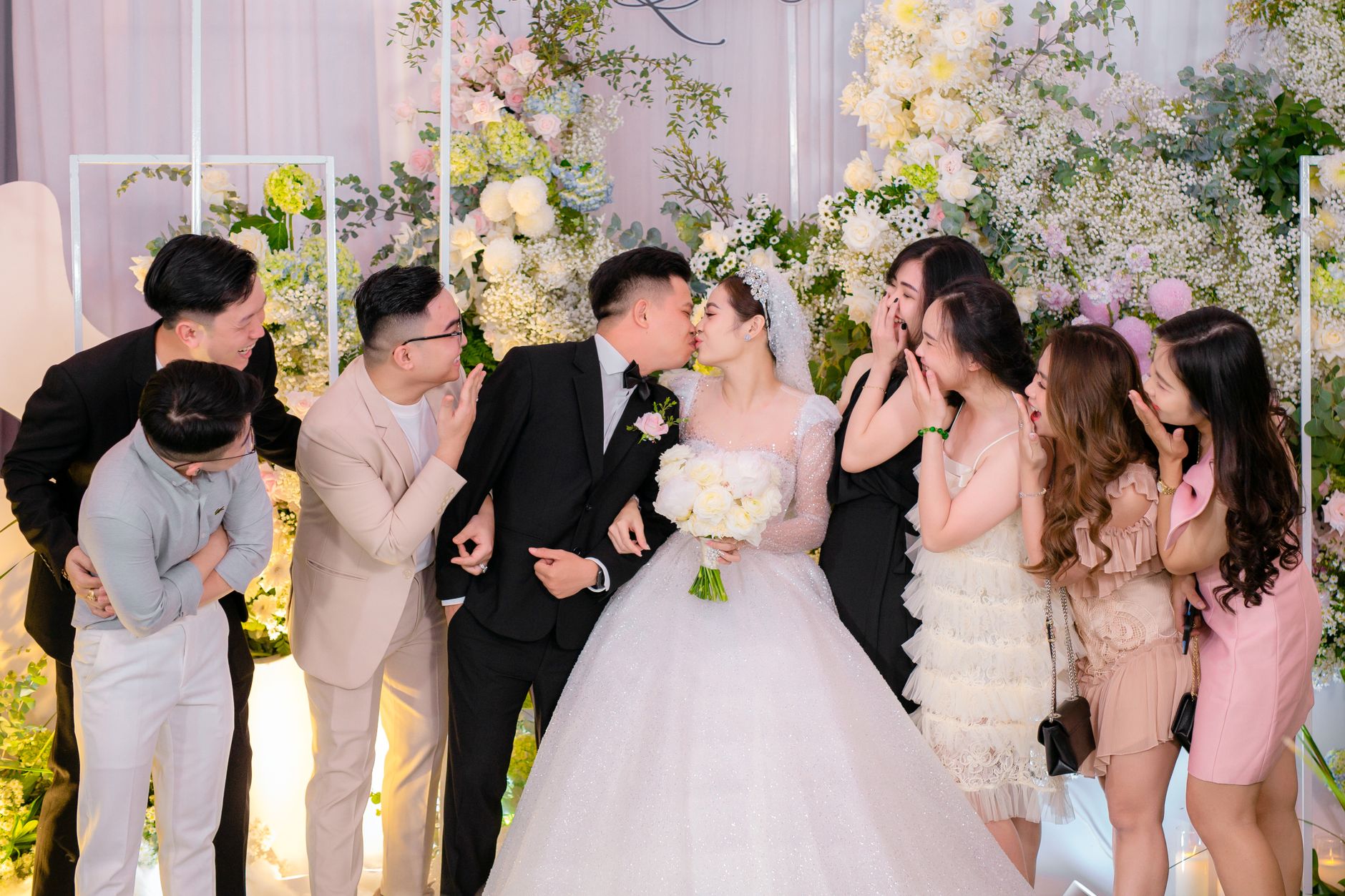 Bride and groom kissing surrounded by joyful friends in a floral setting.