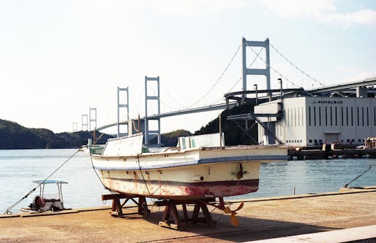 A boat rests at a Japanese harbor with a suspension bridge in the background, under a clear sky.