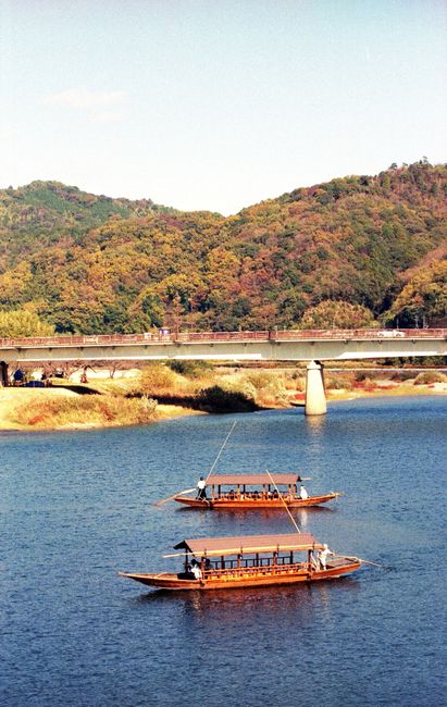 Elegant traditional wooden boats glide on a tranquil Japanese river surrounded by lush hills.