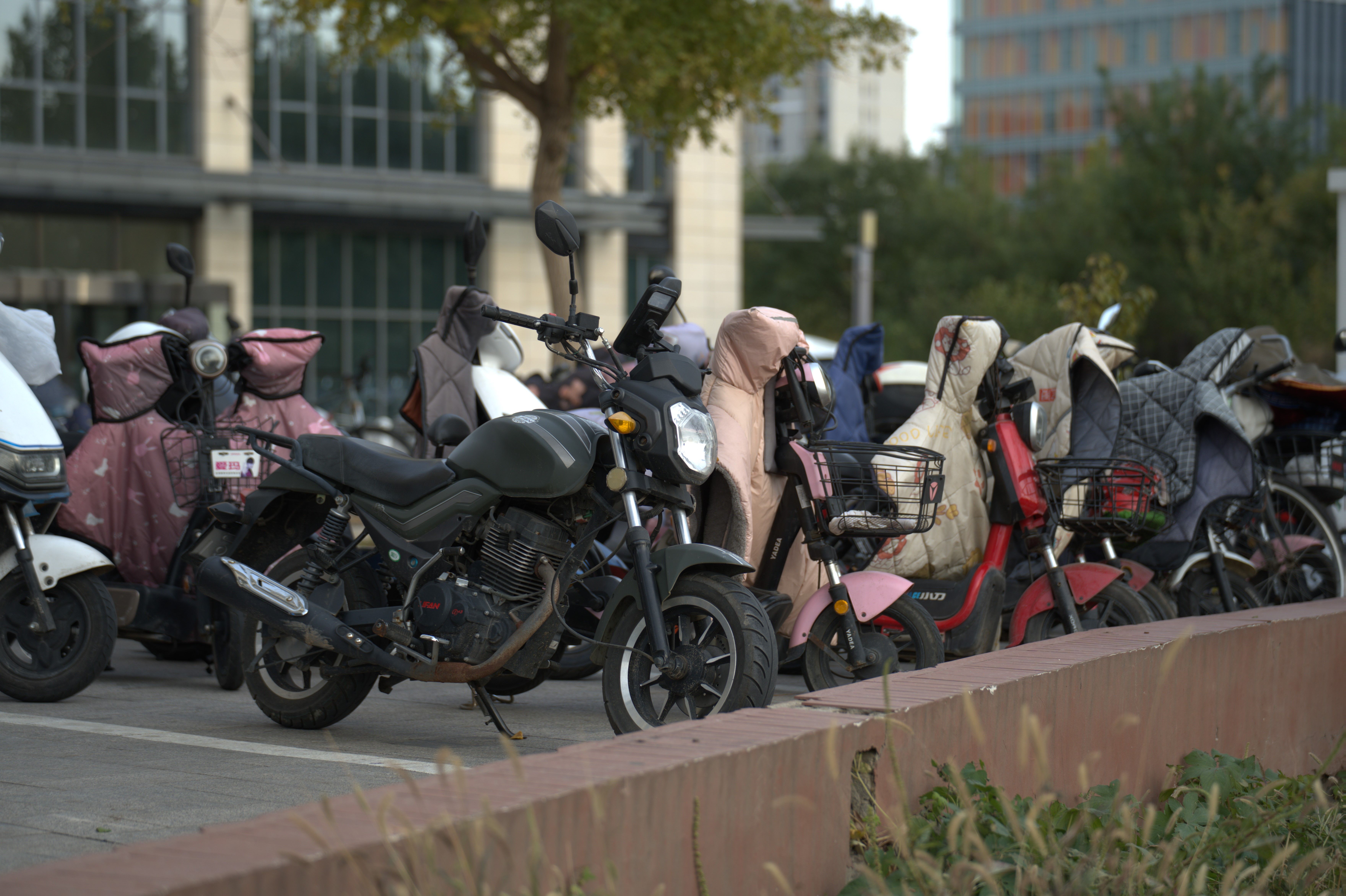 A row of parked scooters and motorcycles in an urban outdoor setting with protective covers.