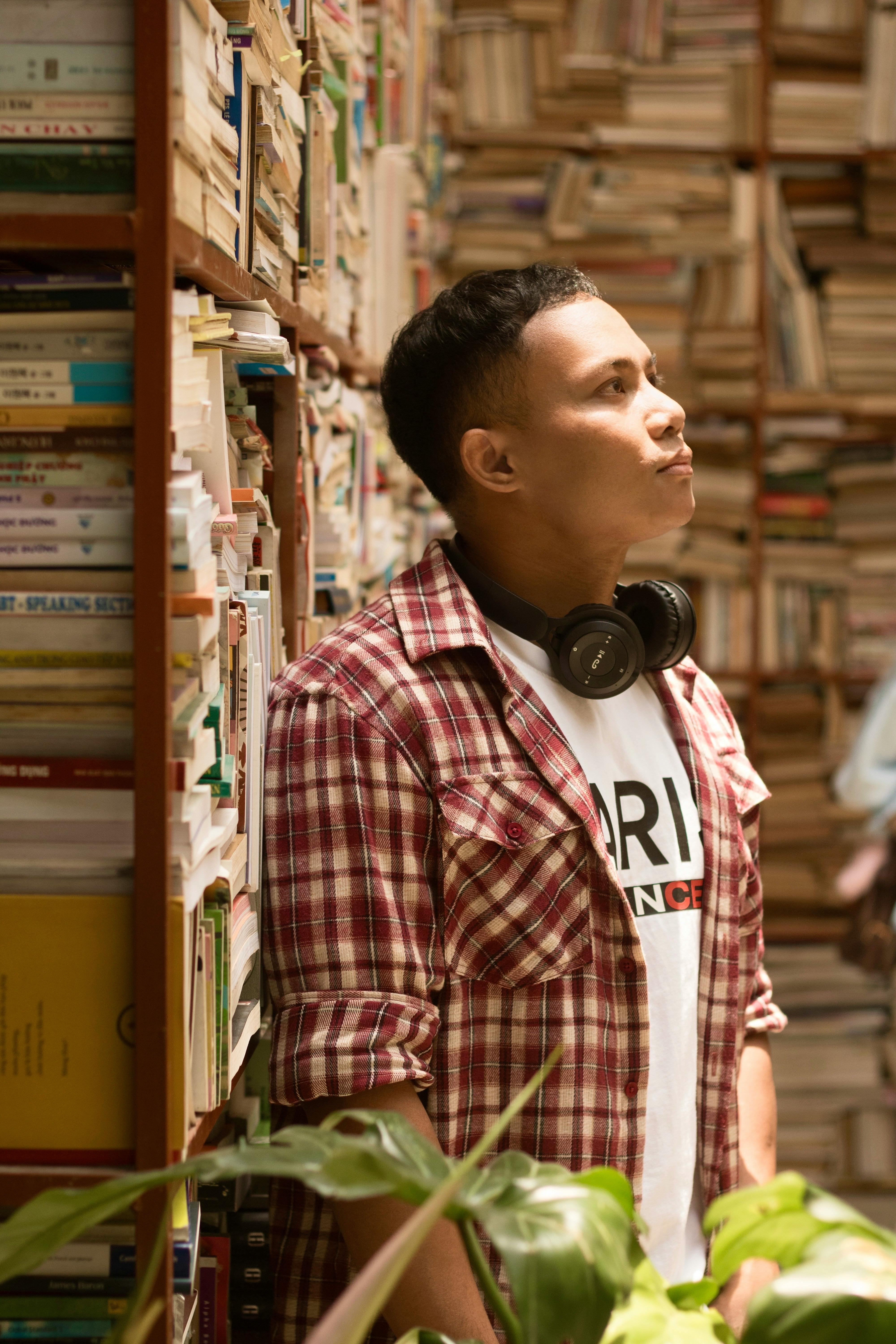 A young man with headphones leans against a bookshelf in a cozy library, deep in thought.