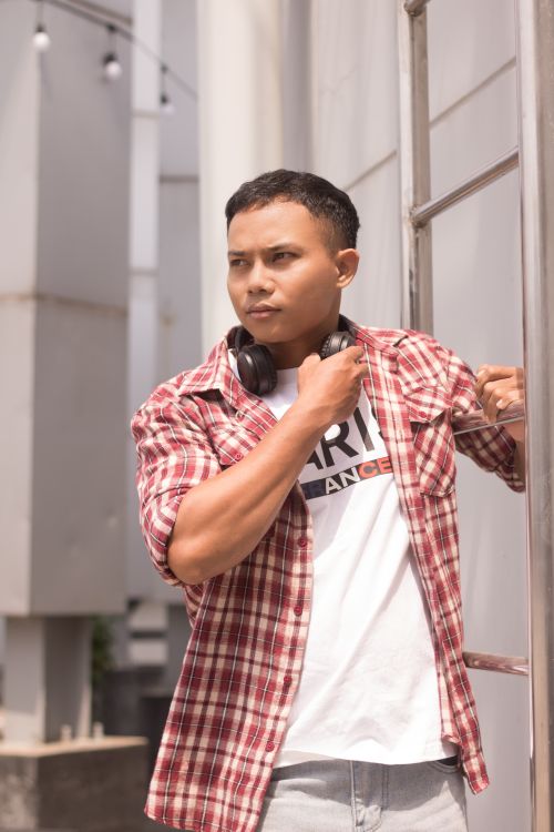 Free A young man in a plaid shirt with headphones poses thoughtfully outdoors. Stock Photo