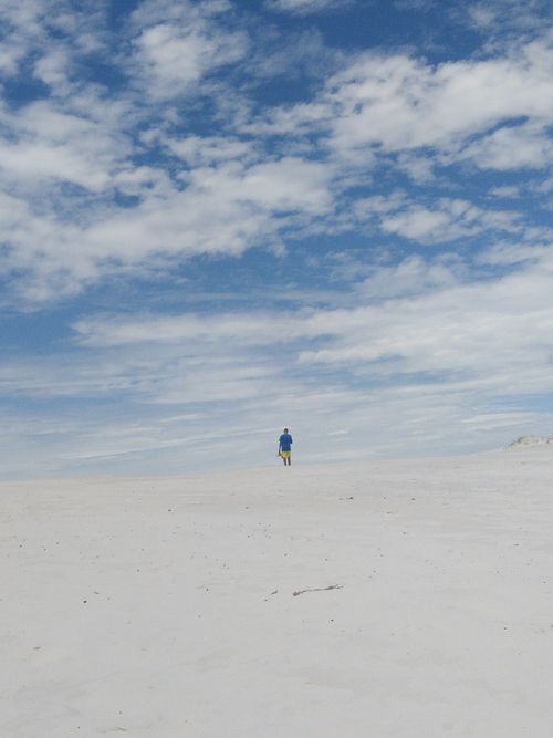 Free A solitary person walks across a vast sandy desert under a bright, cloud-filled sky. Stock Photo