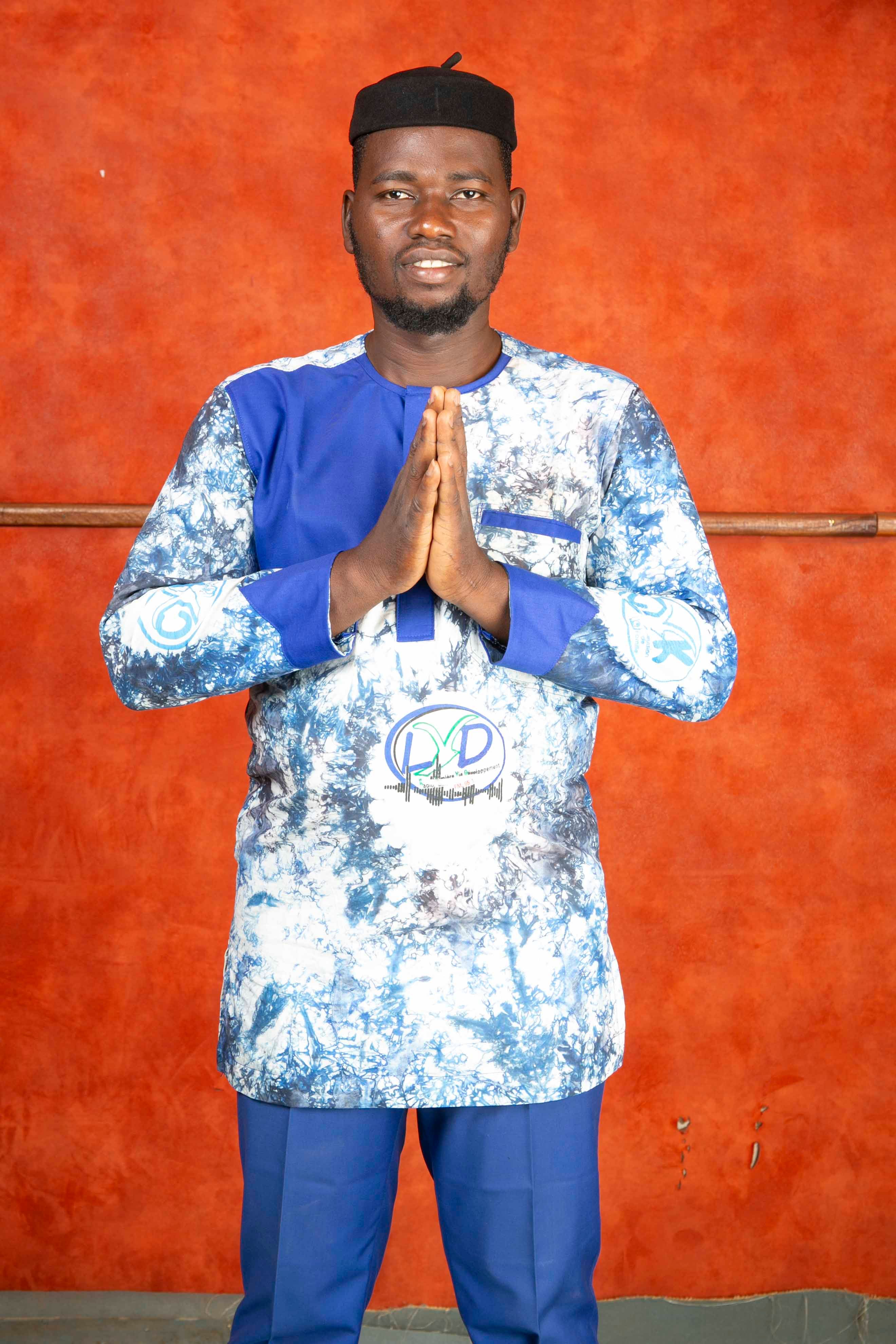 Man standing with hands joined, wearing traditional blue attire and cap against a red background.