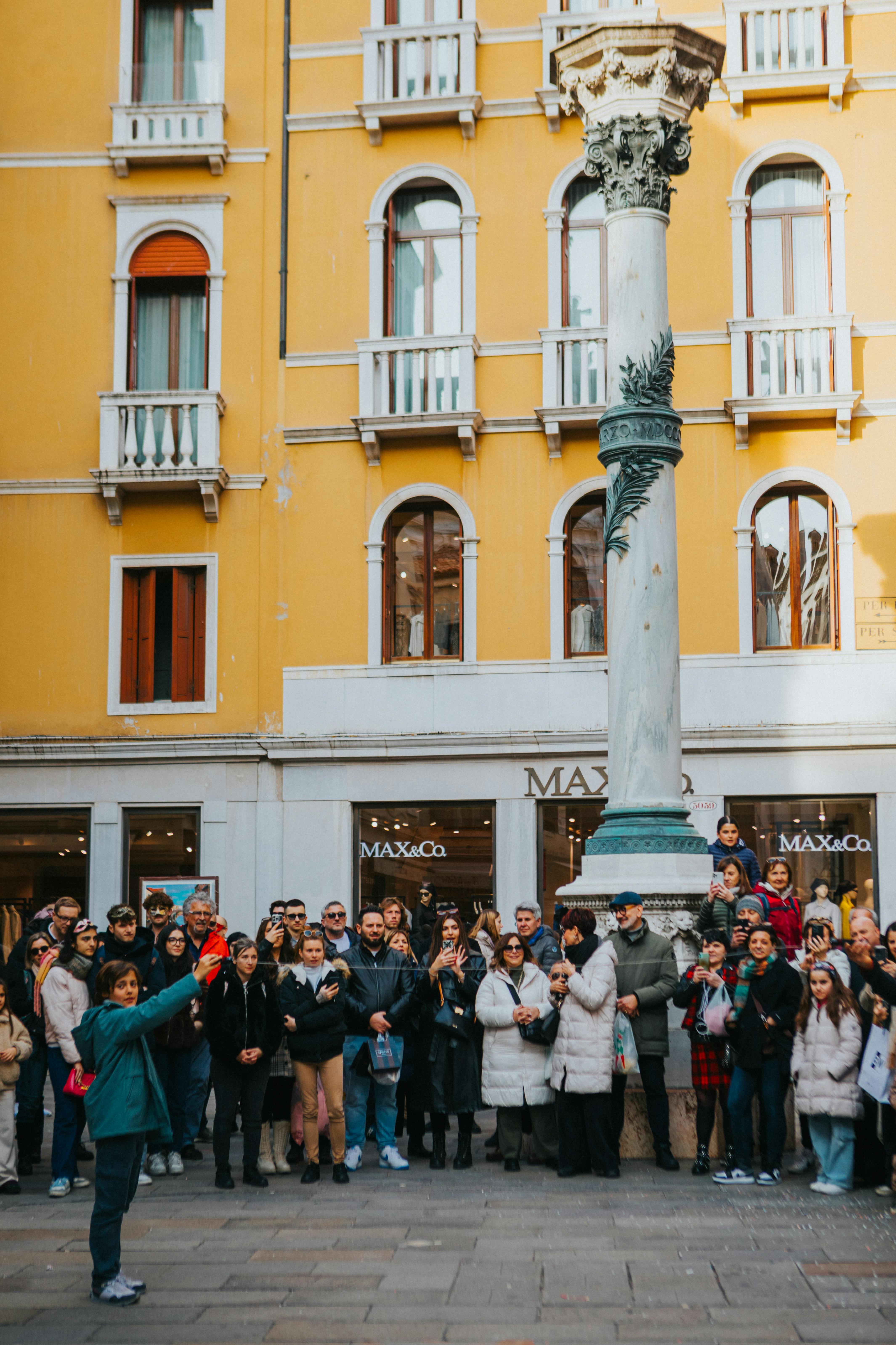A diverse crowd gathers around a historic column in a vibrant urban setting with a yellow building.