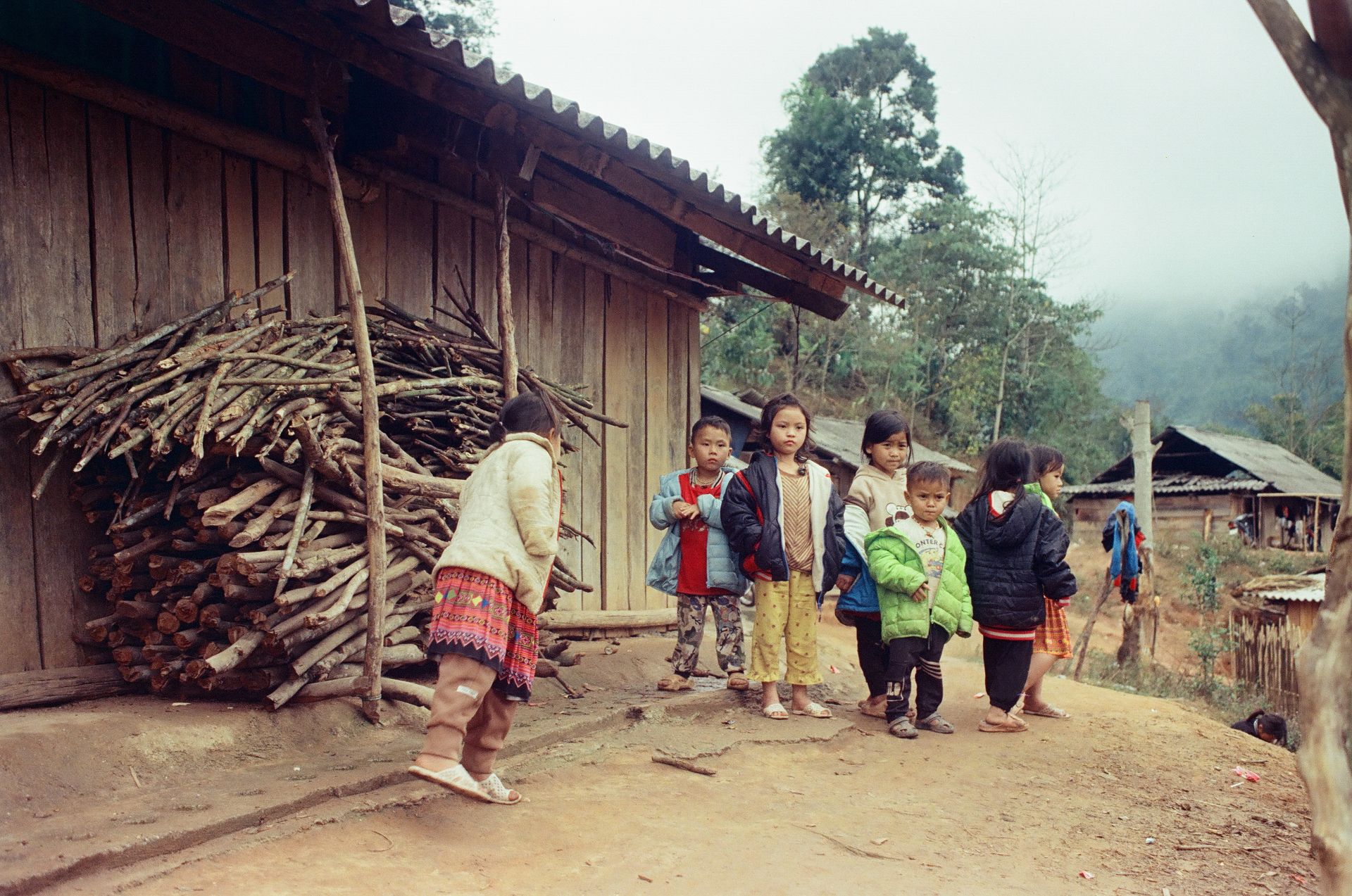 Group of children standing near wooden homes in a rural village setting.