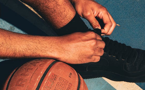 A person ties their basketball shoe on a sunlit outdoor court beside an official game ball.