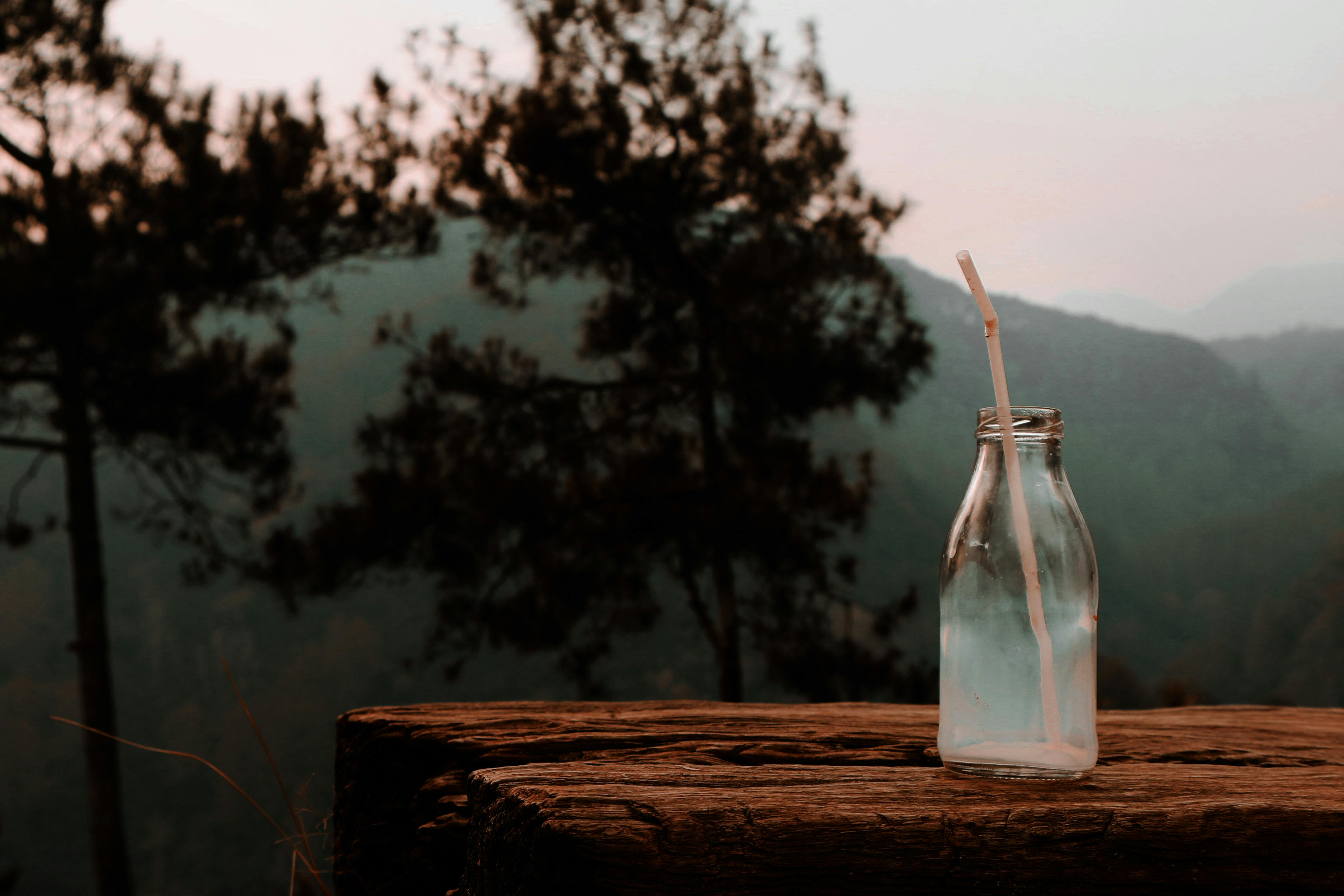 木桌上放着一个玻璃瓶，瓶上插着一根吸管，背景是雾蒙蒙的森林，构成了一幅宁静的场景
