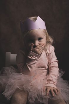 Charming portrait of a child wearing a pink tutu and paper crown, covering mouth playfully.