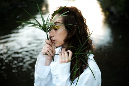 Free A young woman stands elegantly by the waterside holding green leaves, embodying a serene and natural beauty. Stock Photo
