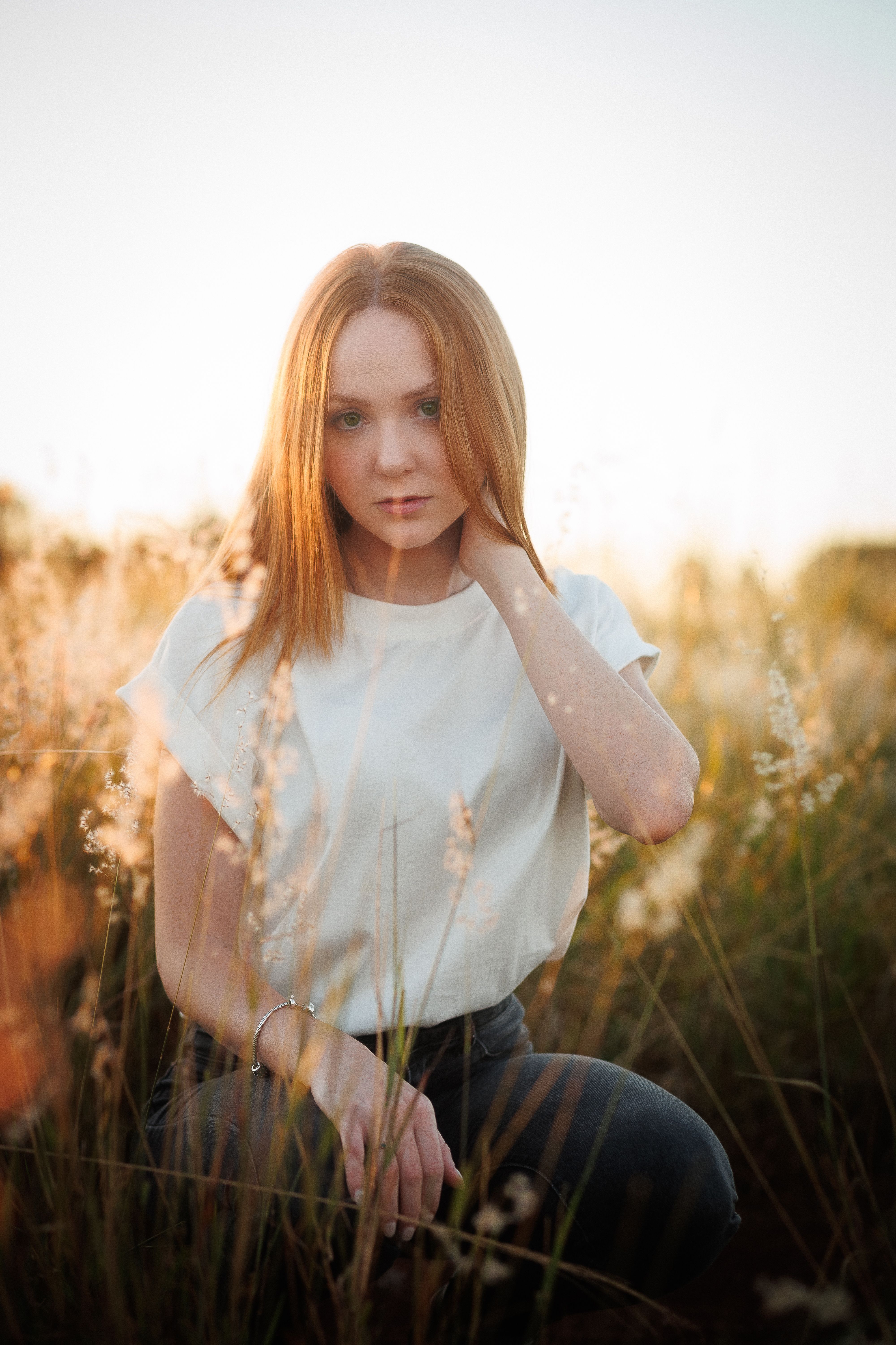 A woman with ginger hair poses in a grassy field under the warm, glowing sunset.