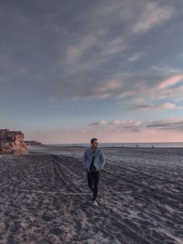 A man strolls along a serene beach at sunset, with dramatic skies and a tranquil ocean backdrop.