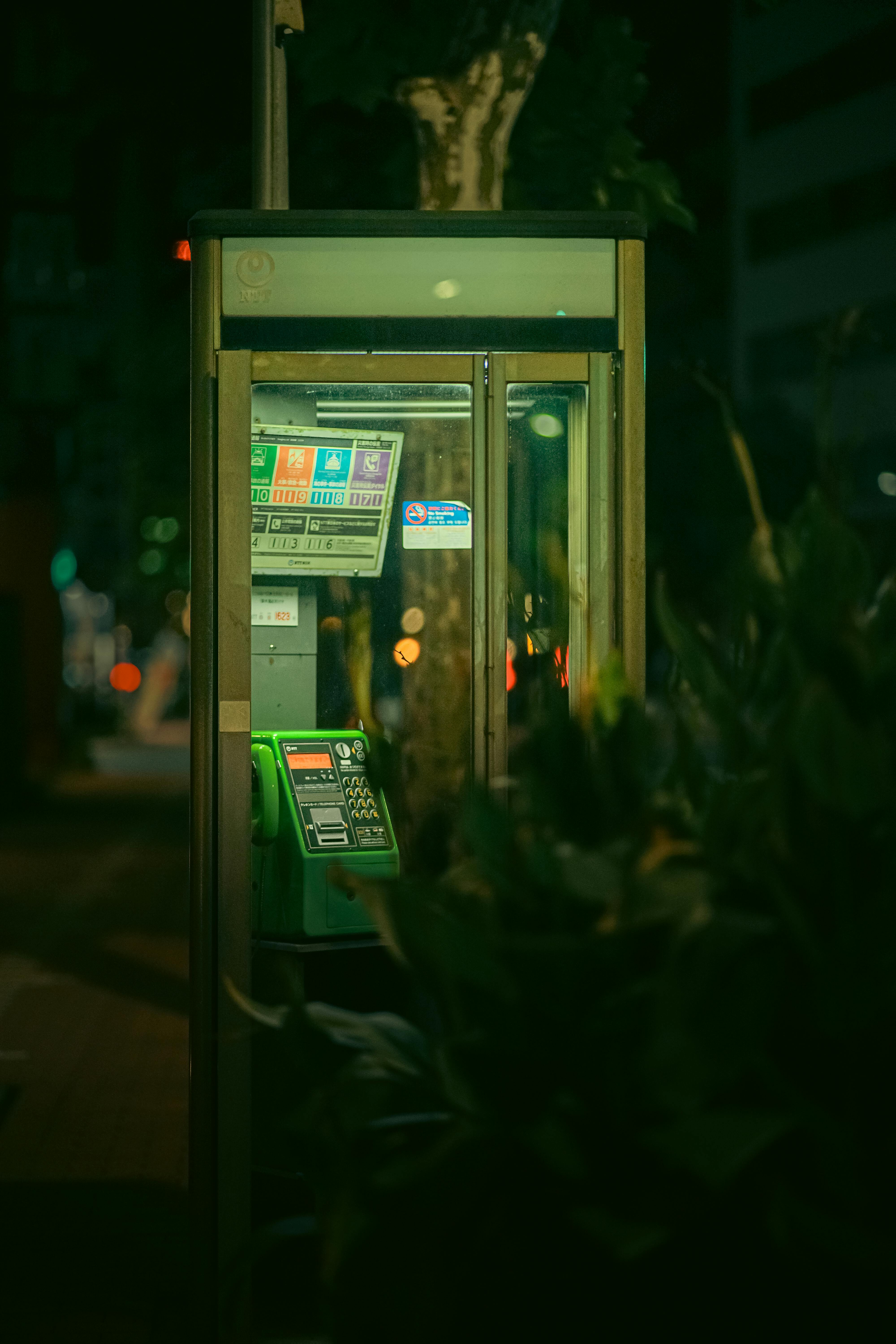 A glowing public phone booth nestled among urban foliage in Shinagawa, Tokyo at night.