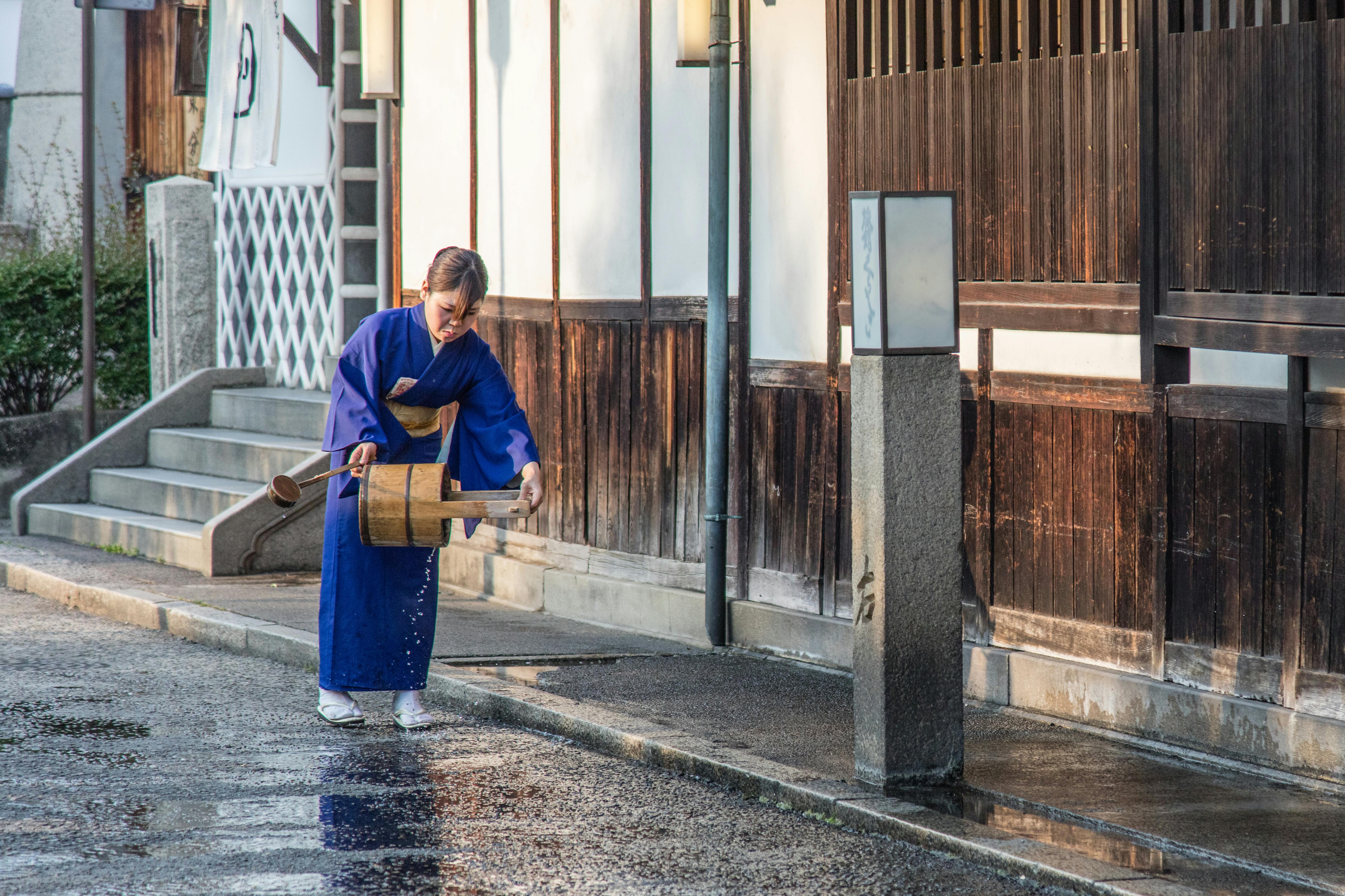Woman in kimono watering street in Kurashiki, Japan, showcasing traditional culture and architecture.