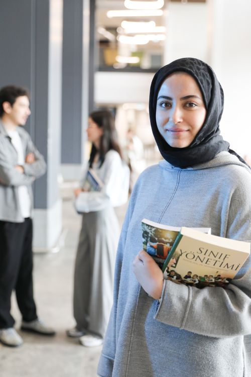 Free Young woman in hijab holding a book while standing in a college hallway, with students in the background. Stock Photo