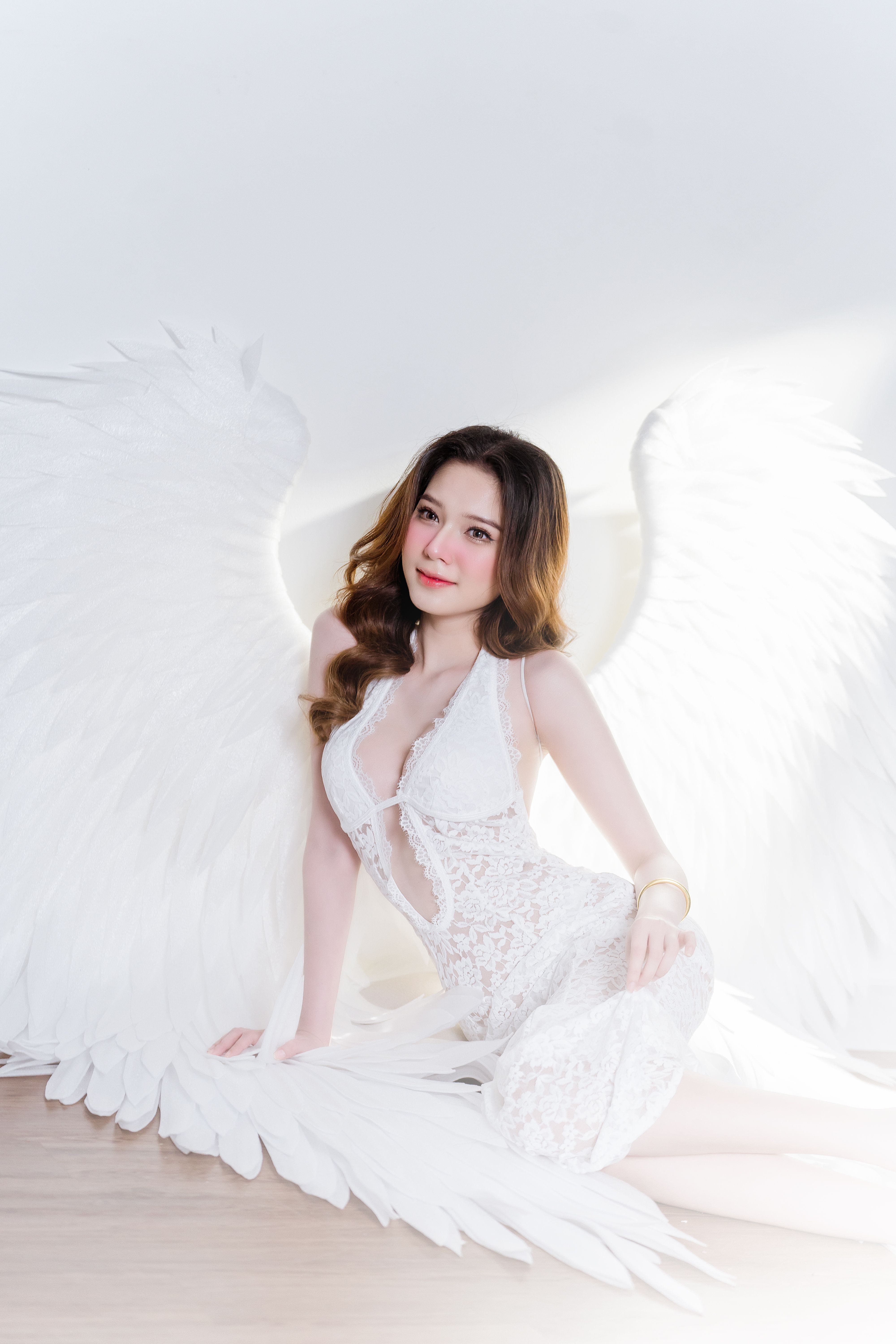 A woman with angel wings in a lace dress poses gracefully in a bright setting.