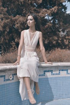 A woman in a light dress sits by an empty pool, showcasing elegance and serenity.
