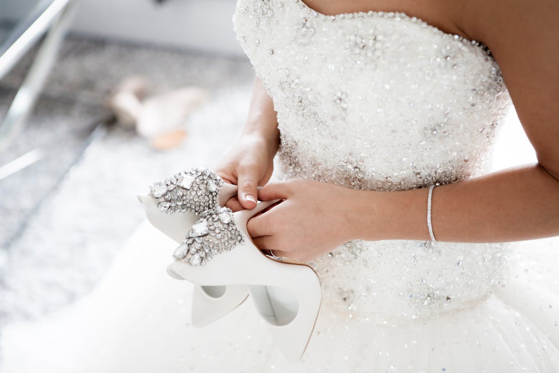 A bride-to-be holds her shoes in her hands as she tries on a wedding gown with a beaded bodice