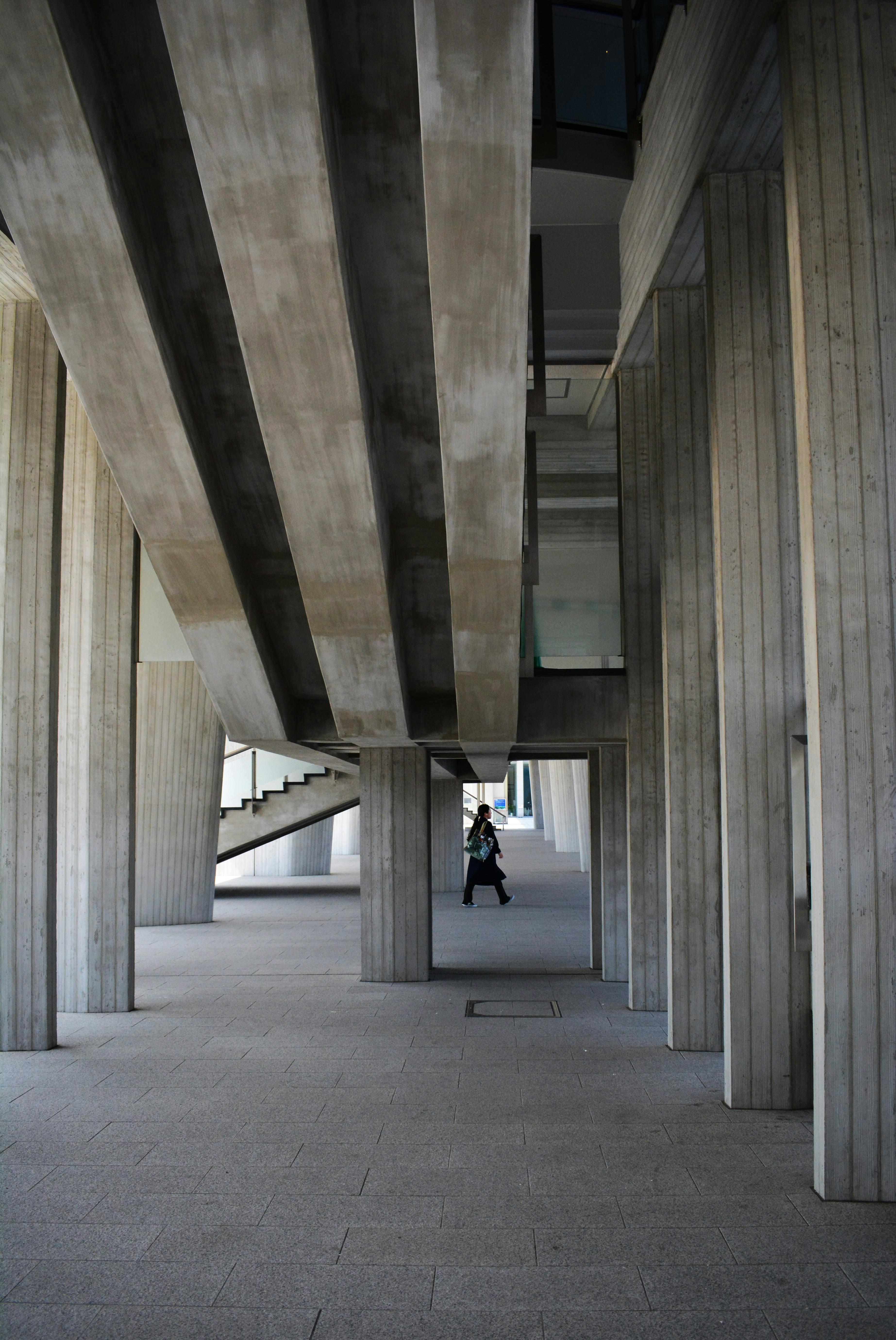 Free A person walking under a modern concrete structure with geometric columns and stairs. Stock Photo