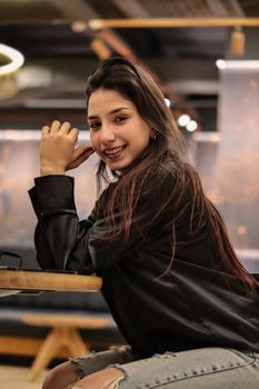 A young woman in a black jacket smiles while sitting indoors, exuding a casual and relaxed vibe.