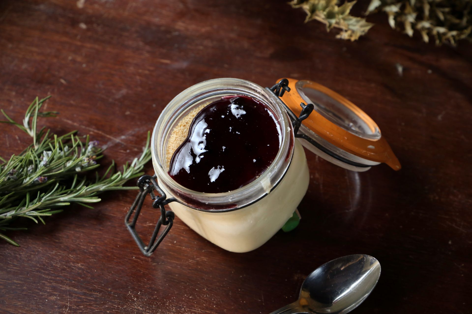 Mason jar with custard and berry jam on wooden surface with rosemary.