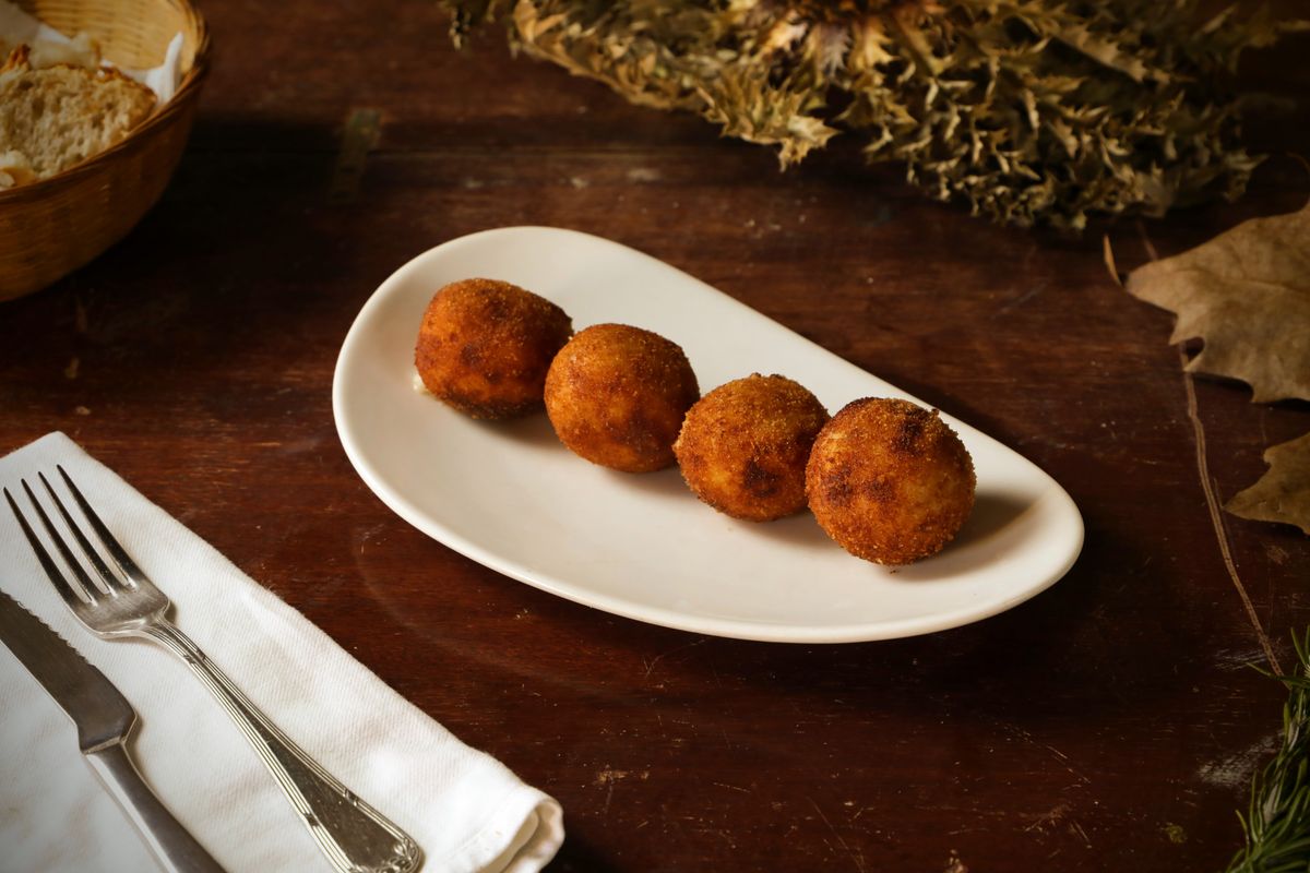 Four golden round arancini on an oval white plate with cutlery on a wooden table