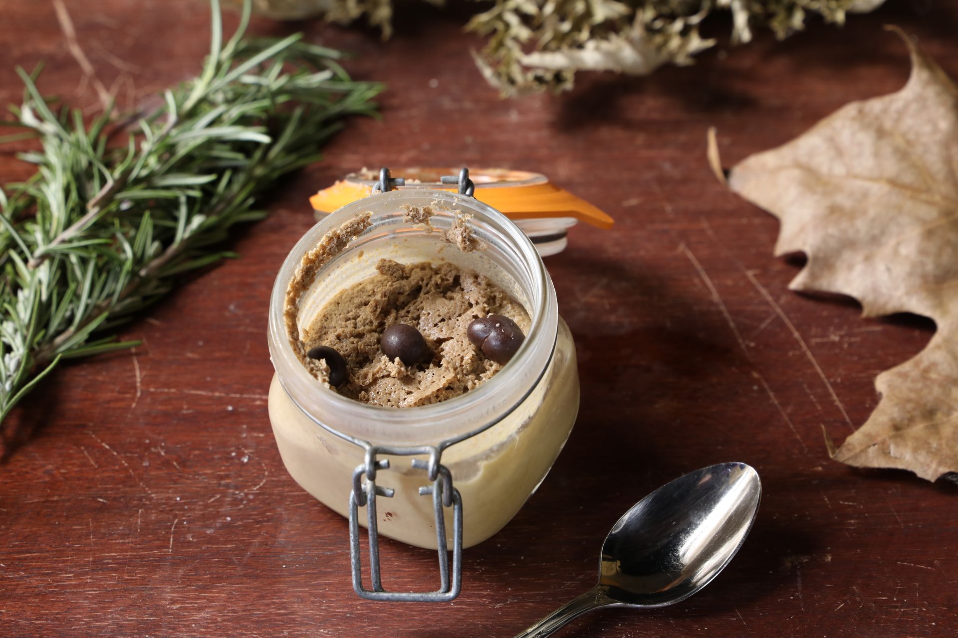 A gourmet spread in a jar with rosemary and olives on a wooden table.