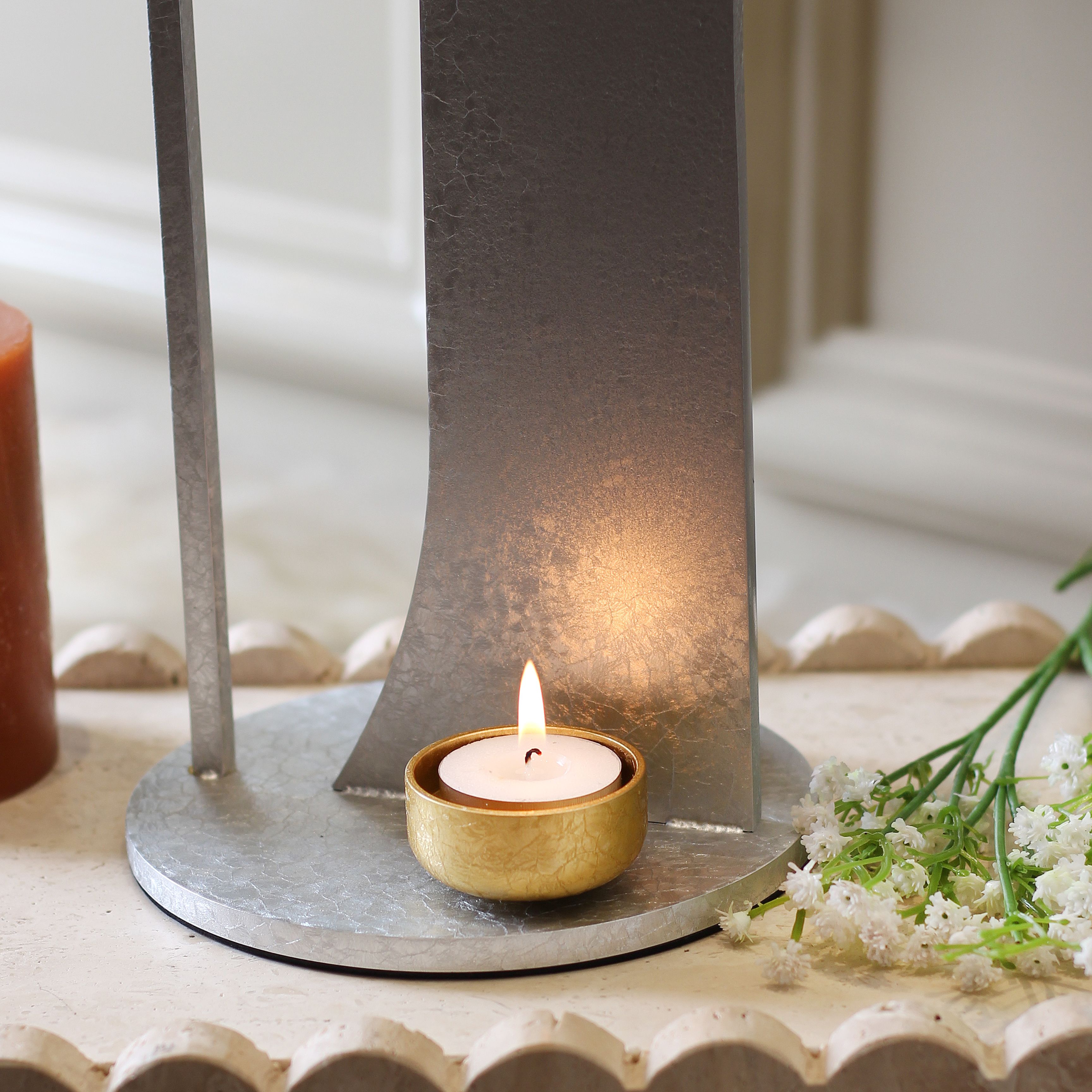 A small candle with golden holder emits a warm glow against a modern metallic stand.