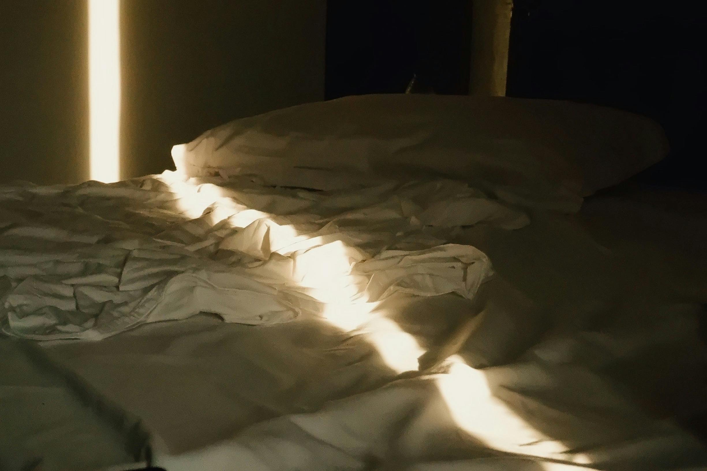 Soft sunlight streaming through window onto an unmade bed, creating a peaceful morning atmosphere.