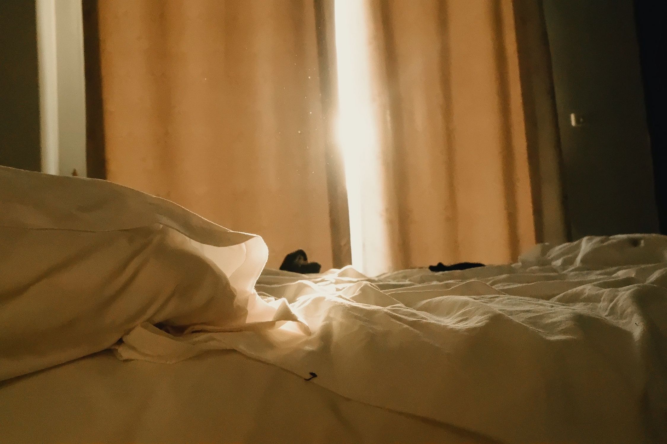 Warm sunlight streaming through curtains onto a cozy, unmade bed in a tranquil room.