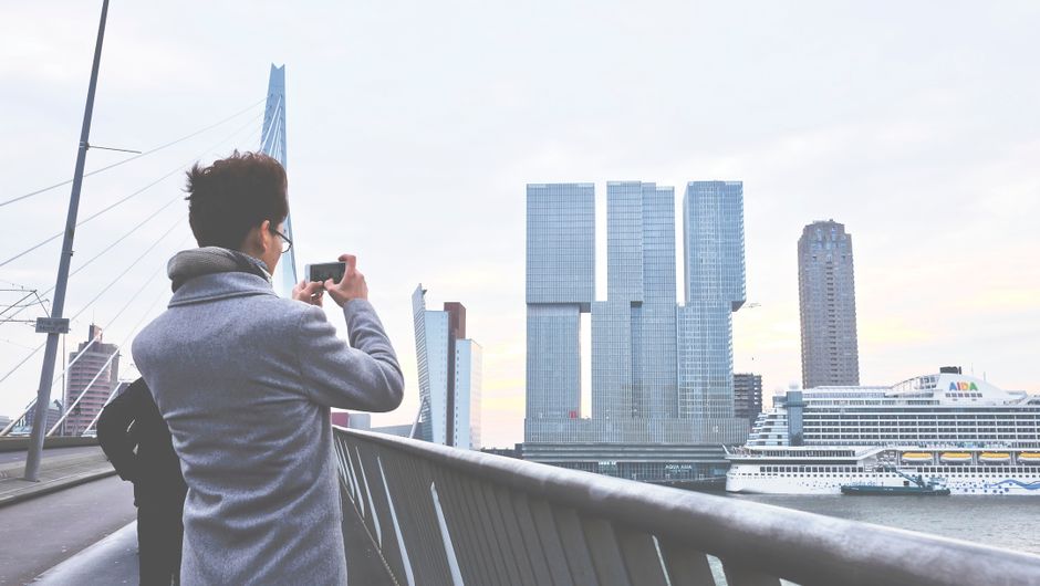 Person photographing Rotterdam's skyline with futuristic architecture and Erasmus Bridge.